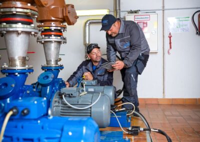Pumpenraum mit blauen Pumpen und Rohrleitungen, zwei Mitarbeiter bei der Arbeit
