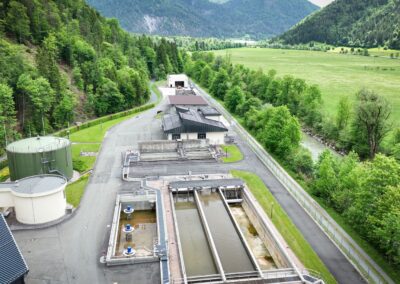 Luftaufnahme der Kläranlage mit langgestreckten Becken, runden Behältern und bewaldetem Hang