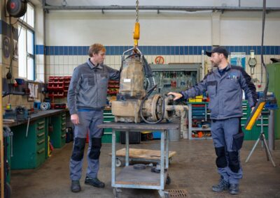 Zwei Mitarbeiter heben eine Pumpe mit dem Kran in der Werkstatt